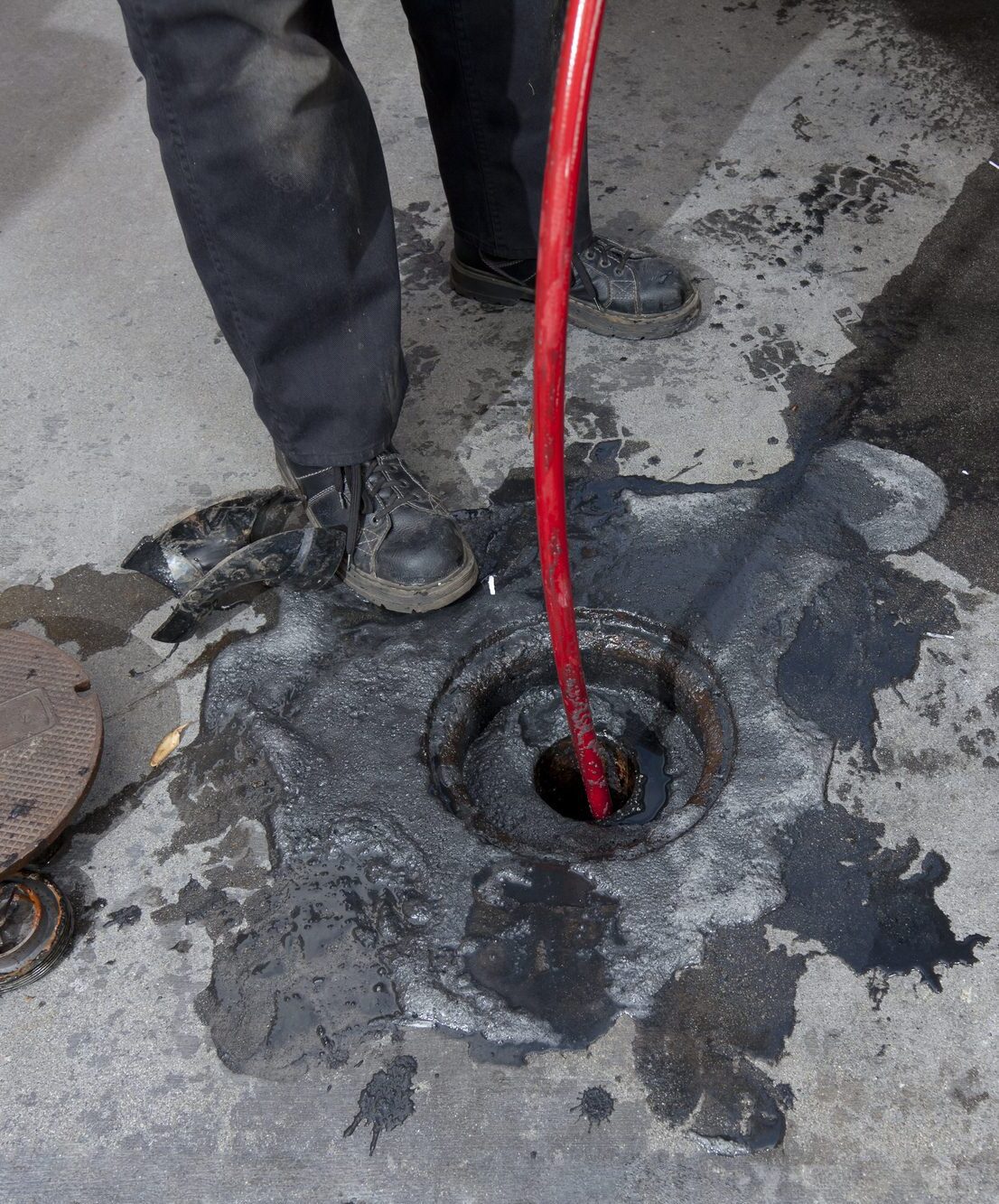 Commercial plumbers hydro line jet a pipe at a large restaurant. Grease can be seen on the pavement surrounding the pipe.